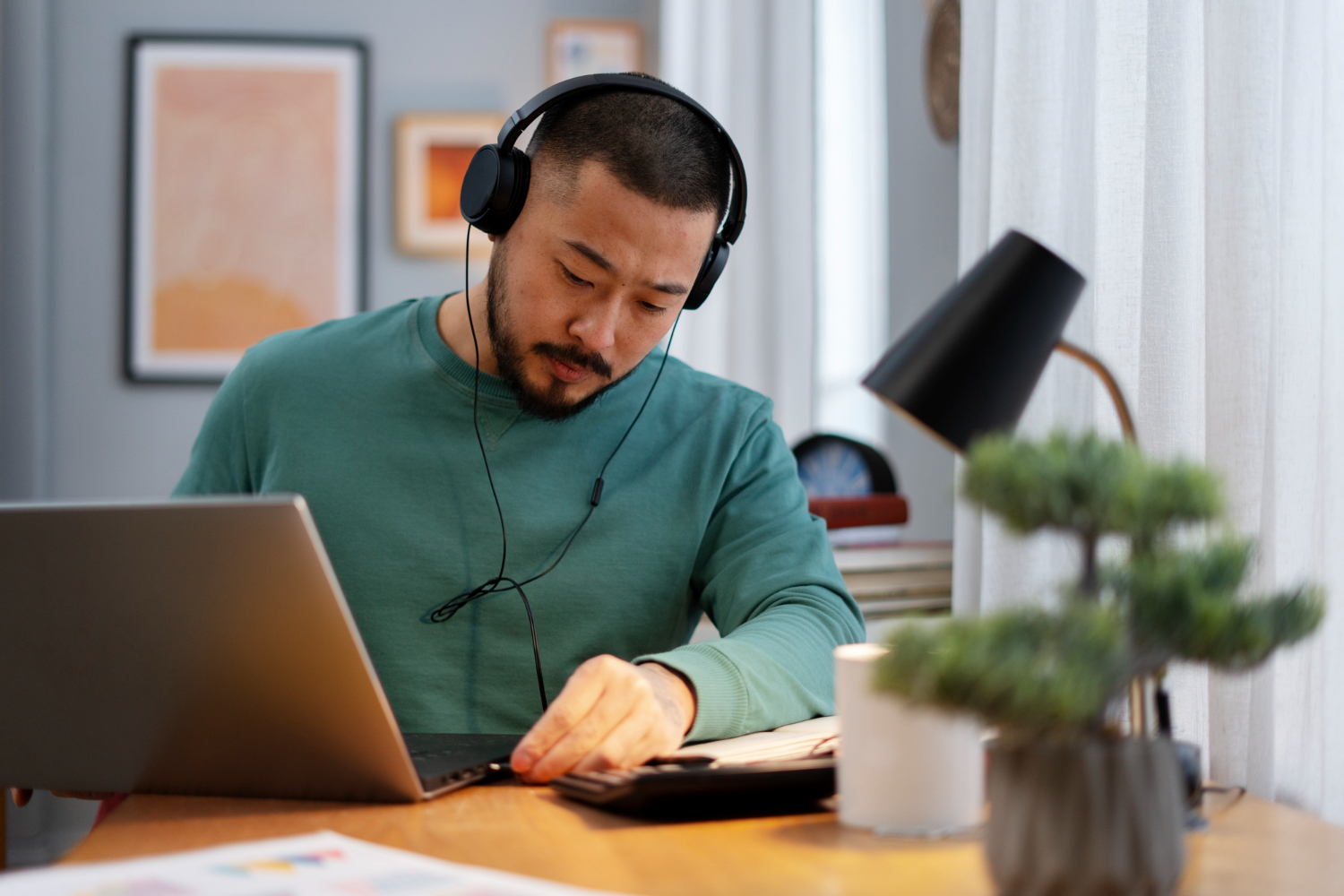 Un homme portant un casque audio et un pull vert travaille avec concentration sur son ordinateur portable à son bureau à domicile