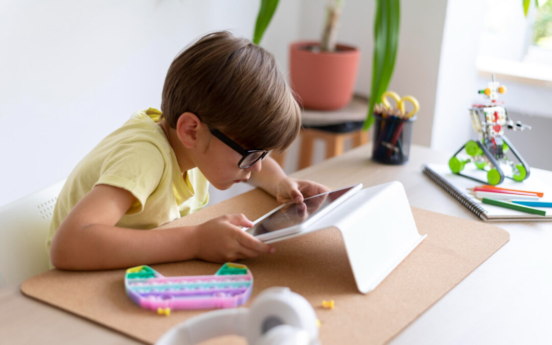 Un enfant assis à une table, regardant une tablette avec concentration. Autour de lui des cahiers ouverts