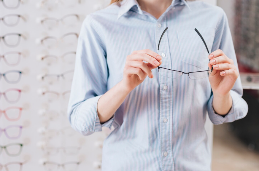 Une personne tient une paire de lunettes de vue devant un présentoir de montures chez un opticien