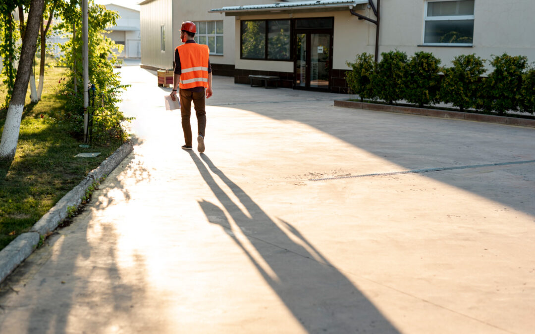 Un ouvrier portant un gilet orange de haute visibilité et un casque de chantier rouge marche de dos sur une grande allée extérieure en béton