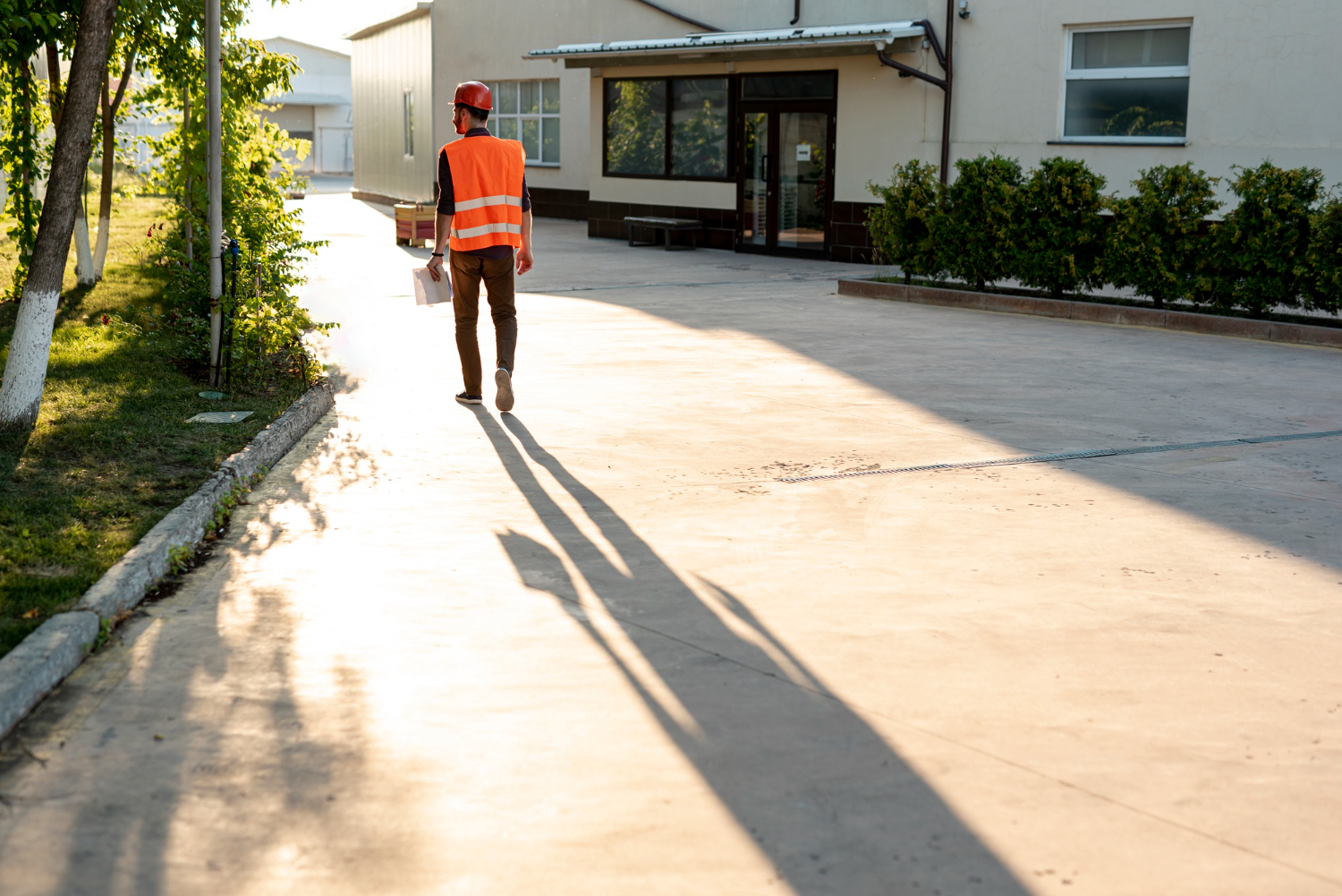 Un ouvrier portant un gilet orange de haute visibilité et un casque de chantier rouge marche de dos sur une grande allée extérieure en béton