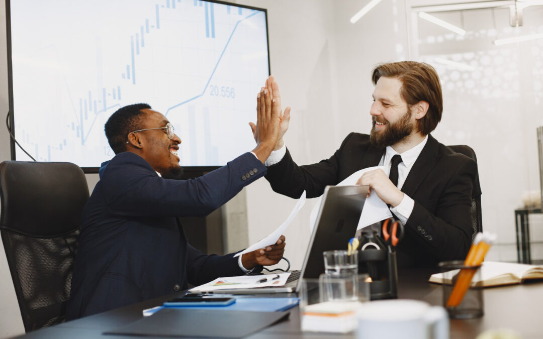 deux hommes d'affaires souriants se tapant dans la main au-dessus d'une table de bureau pour célébrer une réussite professionnelle devant un écran affichant des graphiques.