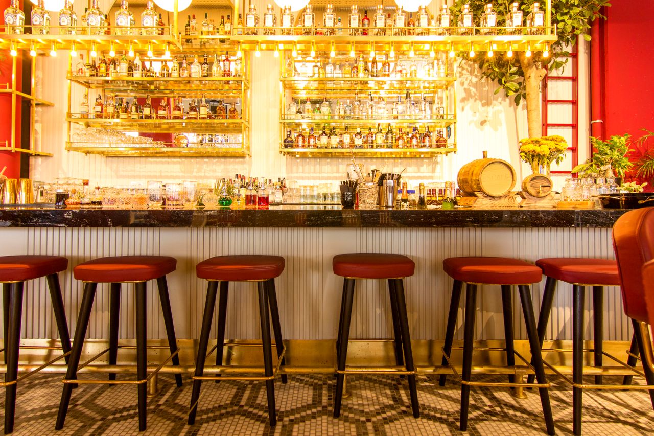 un comptoir de bar chic et moderne aligné de tabourets hauts au siège rouge, surplombé d'étagères dorées et lumineuses chargées de bouteilles de spiritueux