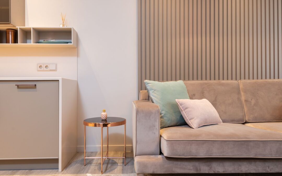 Un salon élégant et moderne aménagé avec un canapé en velours gris, agrémenté de coussins aux teintes douces, à côté d'une petite table basse cuivrée et d'un pan de mur en tasseaux de bois.