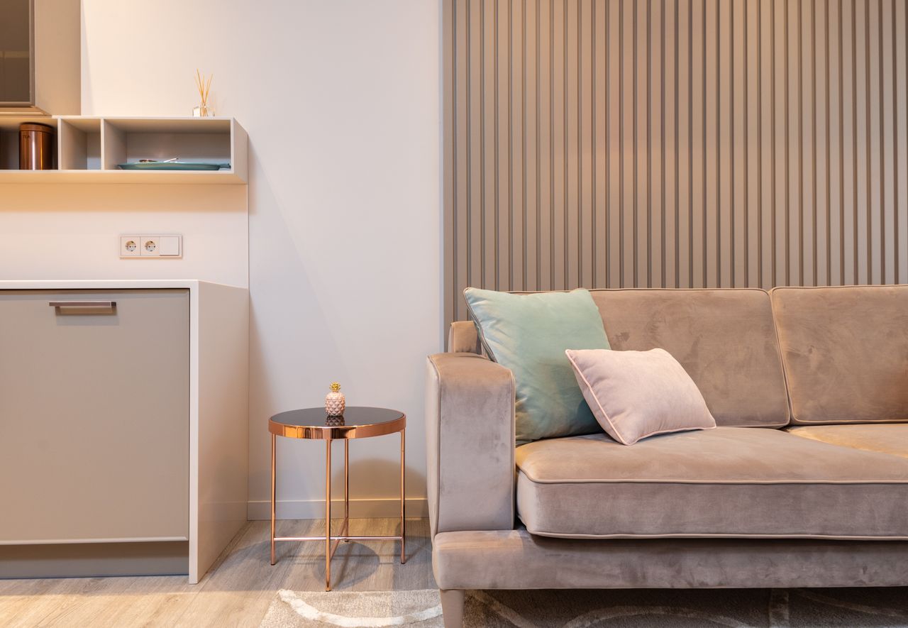 Un salon élégant et moderne aménagé avec un canapé en velours gris, agrémenté de coussins aux teintes douces, à côté d'une petite table basse cuivrée et d'un pan de mur en tasseaux de bois.
