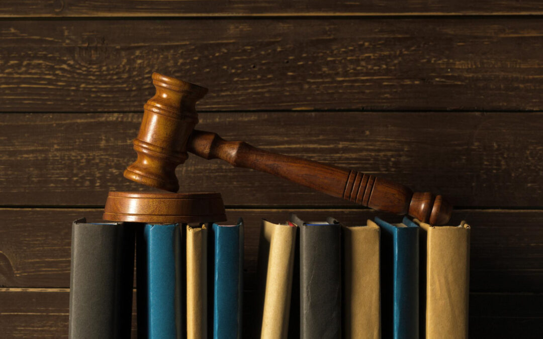 Un marteau de justice en bois repose sur une rangée de livres juridiques alignés à la verticale, le tout posé devant un fond en lattes de bois sombres.