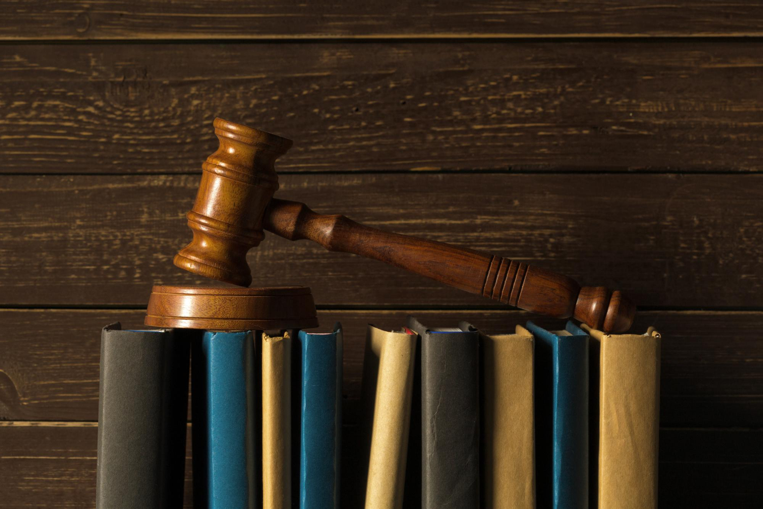 Un marteau de justice en bois repose sur une rangée de livres juridiques alignés à la verticale, le tout posé devant un fond en lattes de bois sombres.